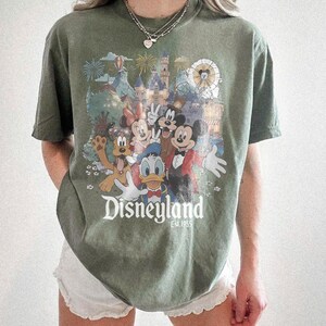 Puede incluir: Camiseta verde oliva con un gr&aacute;fico colorido de Disneyland con Mickey Mouse, Minnie Mouse, Donald Duck y Goofy. El gr&aacute;fico incluye el logotipo de Disneyland y el a&ntilde;o 1955. La camiseta tiene mangas cortas y un corte holgado.