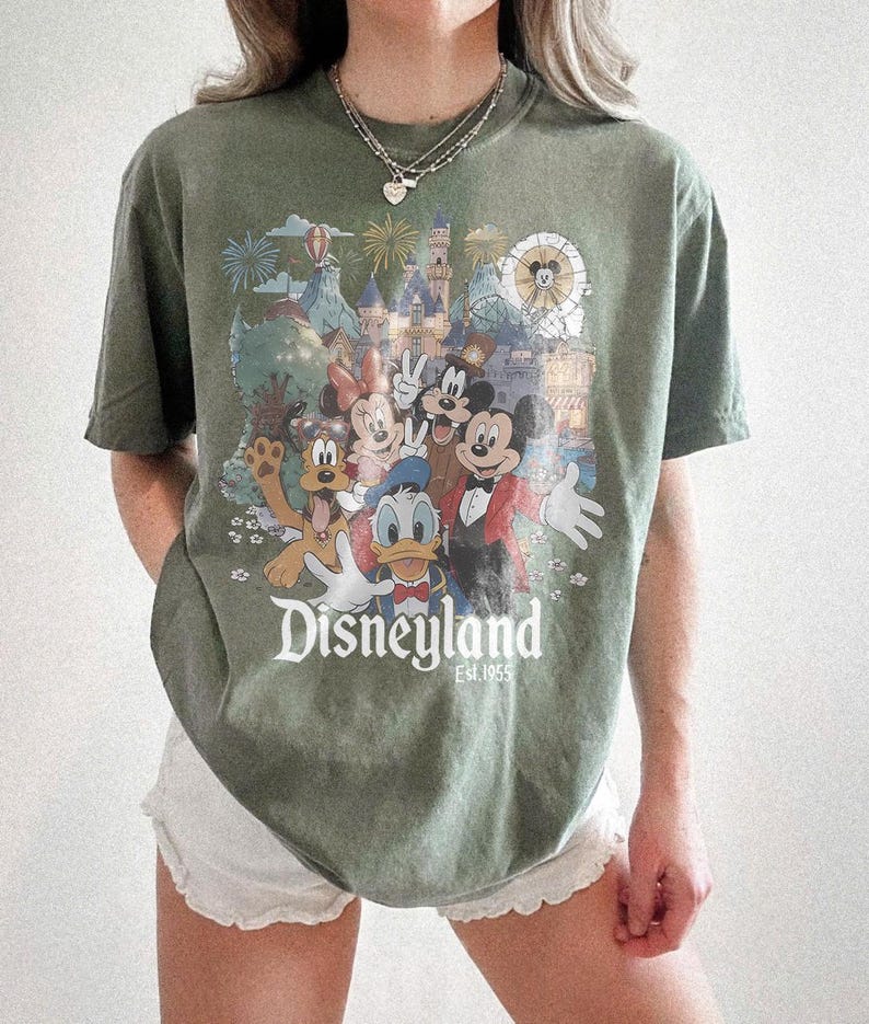 Puede incluir: Camiseta verde oliva con un gr&aacute;fico colorido de Disneyland con Mickey Mouse, Minnie Mouse, Donald Duck y Goofy. El gr&aacute;fico incluye el logotipo de Disneyland y el a&ntilde;o 1955. La camiseta tiene mangas cortas y un corte holgado.