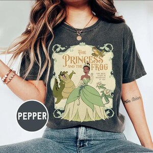 May include: Dark gray t-shirt featuring a graphic from "The Princess and the Frog." The design includes Princess Tiana, an alligator, and a frog, with text that reads "The Princess and the Frog" and "The Real Magic is Believing in You."