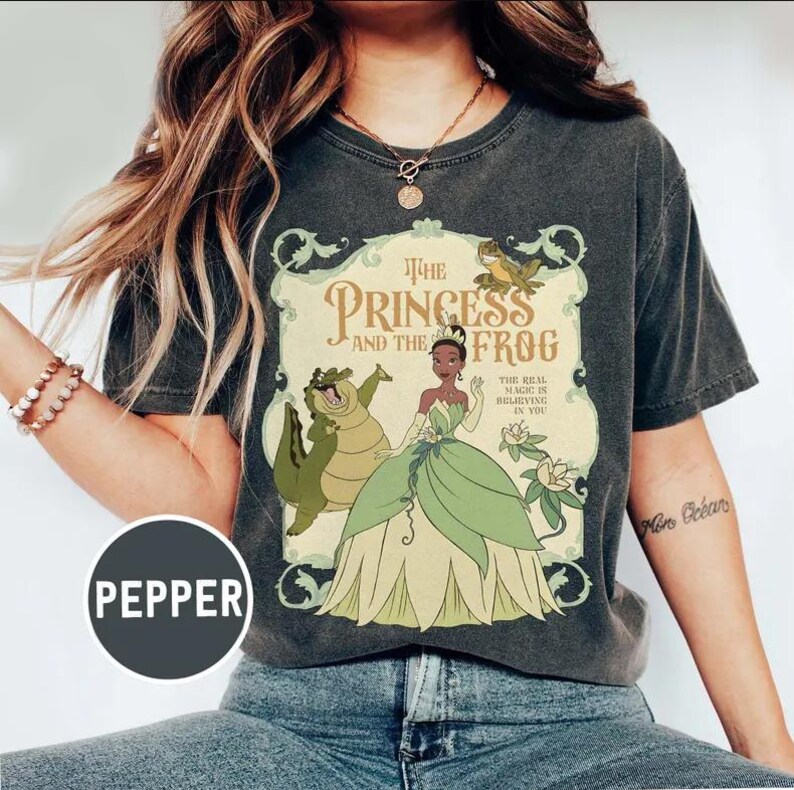 May include: Dark gray t-shirt featuring a graphic from "The Princess and the Frog." The design includes Princess Tiana, an alligator, and a frog, with text that reads "The Princess and the Frog" and "The Real Magic is Believing in You."