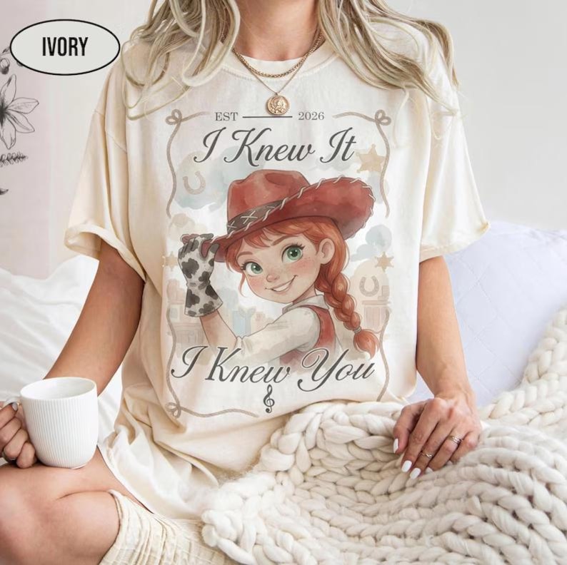 May include: An ivory short-sleeve t-shirt featuring a graphic of a cartoon cowgirl with red hair and a brown hat. The text reads "EST 2026", "I Knew It", and "I Knew You" with a musical note symbol. The design includes a rope border and stars.