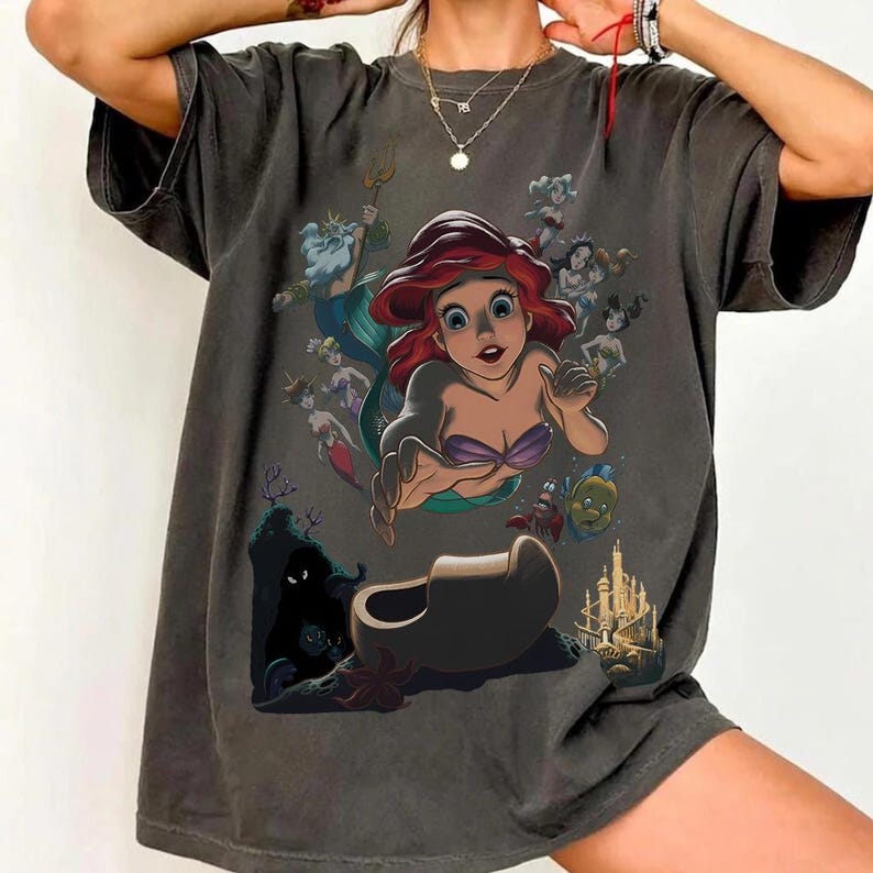 May include: A charcoal gray short-sleeve t-shirt featuring a large graphic print of the character Ariel from The Little Mermaid, surrounded by other characters including King Triton, Flounder, and Sebastian, along with a castle and a dark cave.