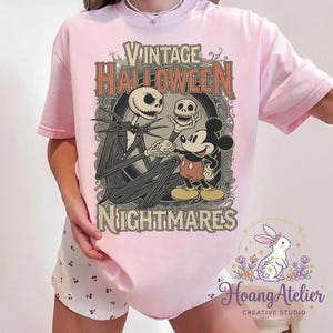 May include: A light pink t-shirt featuring a graphic print with the text "VINTAGE HALLOWEEN NIGHTMARES". The design depicts a tall skeletal figure holding a skull and shaking hands with a cartoon mouse character, framed by ornate scrollwork.