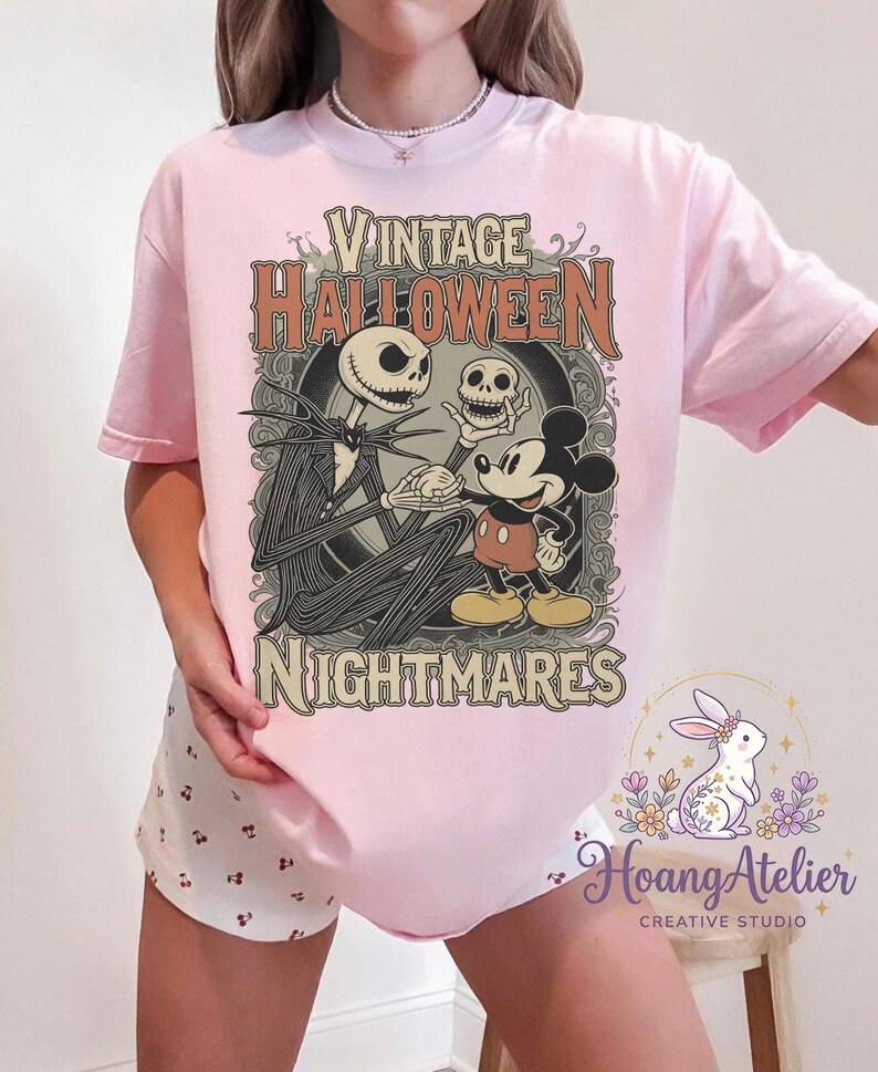 May include: A light pink t-shirt featuring a graphic print with the text "VINTAGE HALLOWEEN NIGHTMARES". The design depicts a tall skeletal figure holding a skull and shaking hands with a cartoon mouse character, framed by ornate scrollwork.