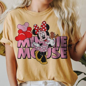 May include: Mustard yellow t-shirt featuring a graphic of Minnie Mouse holding balloons and an ice cream cone. Minnie Mouse is wearing a pink polka dot dress and a red polka dot bow. The text "MINNIE MOUSE" is in pink.