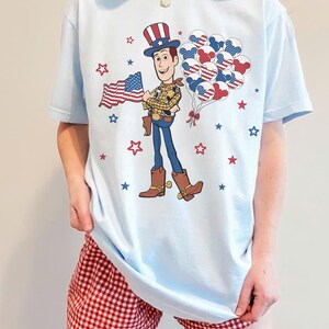 May include: Pale blue t-shirt with a cartoon cowboy graphic holding a US flag and balloons. The cowboy is wearing a hat, boots, and a sheriff's badge. Red and blue stars are scattered around the design. The shirt is worn with red and white checked shorts.