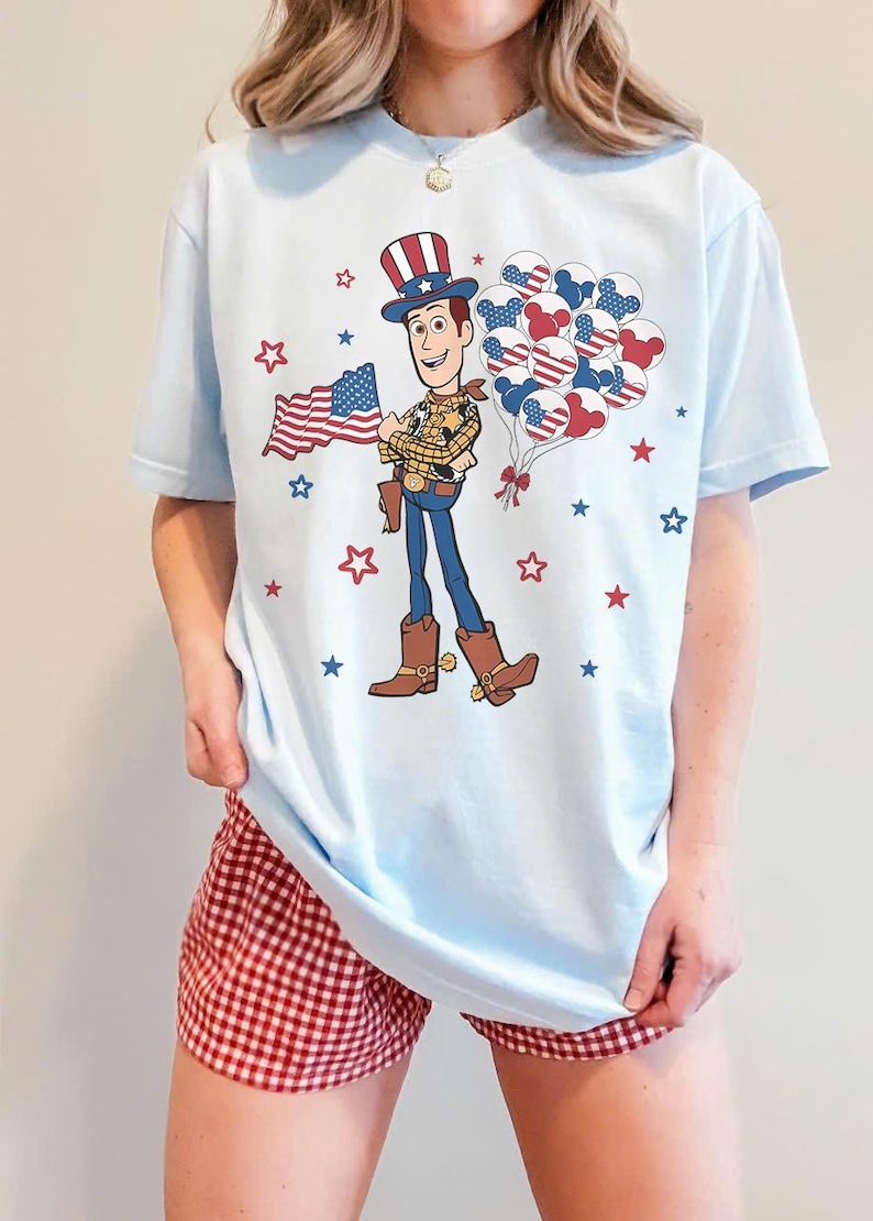 May include: Pale blue t-shirt with a cartoon cowboy graphic holding a US flag and balloons. The cowboy is wearing a hat, boots, and a sheriff's badge. Red and blue stars are scattered around the design. The shirt is worn with red and white checked shorts.