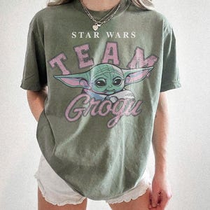 Puede incluir: Una camiseta de manga corta color verde oliva con un estampado gr&aacute;fico de Grogu de Star Wars. El texto dice STAR WARS y TEAM GROGU en una fuente rosa desgastada. La camiseta tiene cuello redondo y un corte holgado de estilo vintage.