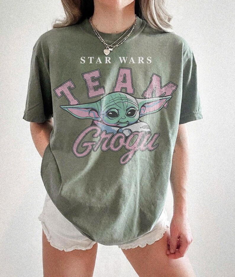 Puede incluir: Una camiseta de manga corta color verde oliva con un estampado gr&aacute;fico de Grogu de Star Wars. El texto dice STAR WARS y TEAM GROGU en una fuente rosa desgastada. La camiseta tiene cuello redondo y un corte holgado de estilo vintage.