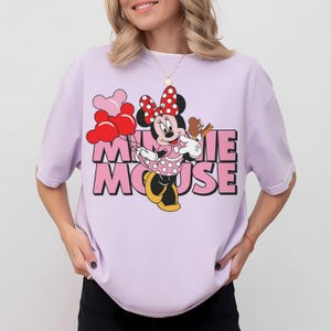 May include: A light purple t-shirt featuring Minnie Mouse holding balloons and a pretzel. The text "MINNIE MOUSE" is printed in pink with a black outline. Minnie Mouse is wearing a polka dot dress and a red bow.