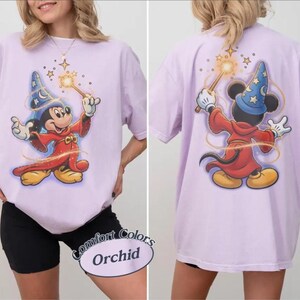 May include: A lilac t-shirt with a cartoon image of Mickey Mouse as a wizard, holding a wand with stars. The back of the shirt also has the same image. The shirt has the words "Comfort Colors Orchid" on the bottom.