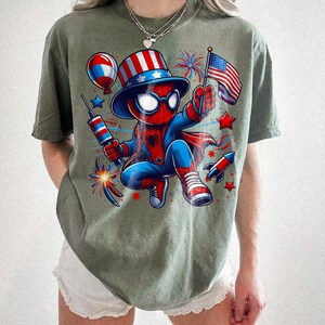 K&ouml;nnte beinhalten: Olivgr&uuml;nes T-Shirt mit einer Comicfigur von Spider-Man in einem patriotischen Outfit. Die Figur h&auml;lt eine kleine amerikanische Flagge und ein Feuerwerk. Das Design umfasst rote, wei&szlig;e und blaue Elemente.