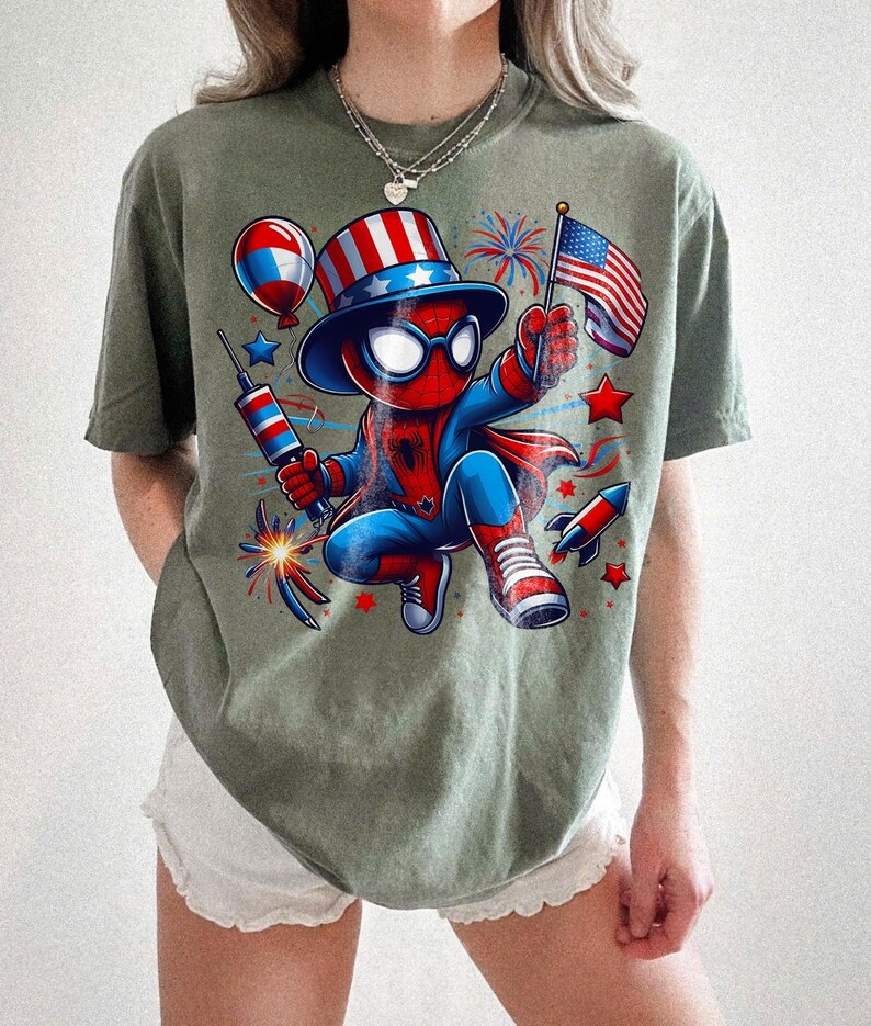 K&ouml;nnte beinhalten: Olivgr&uuml;nes T-Shirt mit einer Comicfigur von Spider-Man in einem patriotischen Outfit. Die Figur h&auml;lt eine kleine amerikanische Flagge und ein Feuerwerk. Das Design umfasst rote, wei&szlig;e und blaue Elemente.
