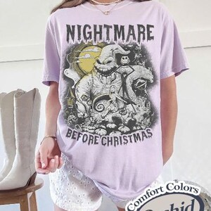 May include: A light purple short-sleeve t-shirt featuring a graphic print of characters from The Nightmare Before Christmas, including Oogie Boogie, Jack Skellington, and Sally, with the text NIGHTMARE BEFORE CHRISTMAS printed in black.