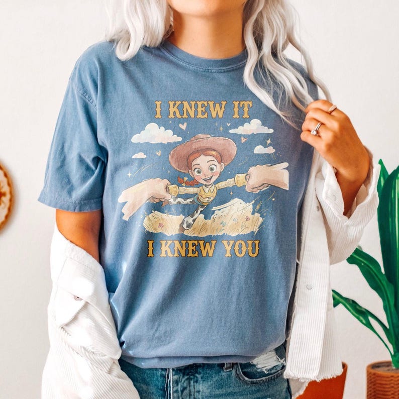 May include: A blue short-sleeved t-shirt featuring a graphic of the character Jessie from Toy Story. The design includes the text I KNEW IT above the character and I KNEW YOU below, with clouds, stars, and two hands reaching toward the character.