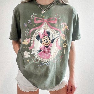 May include: An olive green short-sleeve t-shirt featuring a graphic print of Minnie Mouse in a pink polka-dot dress and bow. The design includes a pearl-bordered oval frame, a large pink ribbon, small stars, and floral accents in soft pastel tones.