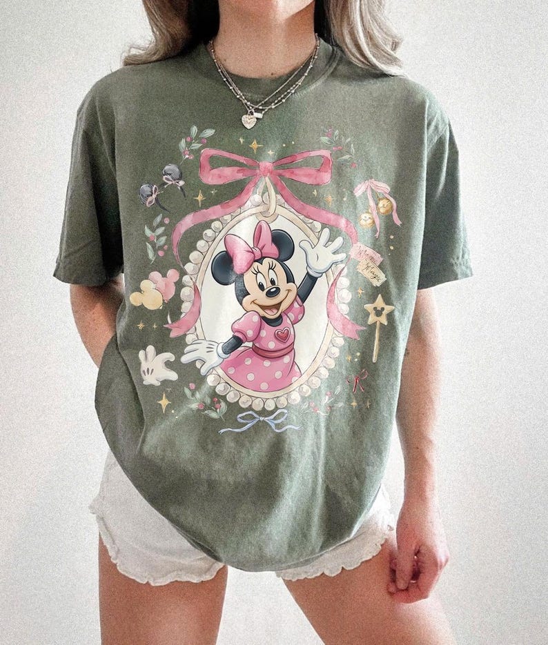 May include: An olive green short-sleeve t-shirt featuring a graphic print of Minnie Mouse in a pink polka-dot dress and bow. The design includes a pearl-bordered oval frame, a large pink ribbon, small stars, and floral accents in soft pastel tones.