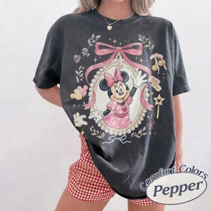 May include: A charcoal gray short-sleeve t-shirt featuring a graphic of Minnie Mouse in a pink polka-dot dress inside an ornate oval frame. The design includes a large pink bow, floral accents, small stars, and a star-shaped wand. The shirt has a crew neck.