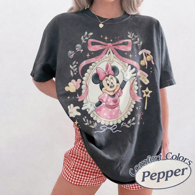 May include: A charcoal gray short-sleeve t-shirt featuring a graphic of Minnie Mouse in a pink polka-dot dress inside an ornate oval frame. The design includes a large pink bow, floral accents, small stars, and a star-shaped wand. The shirt has a crew neck.