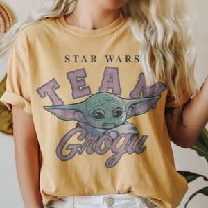 Puede incluir: Una camiseta de manga corta color amarillo mostaza con un estampado gr&aacute;fico del personaje Grogu de Star Wars. El texto STAR WARS aparece en letras negras sobre la palabra TEAM en morado y Grogu en fuente cursiva morada bajo el personaje.