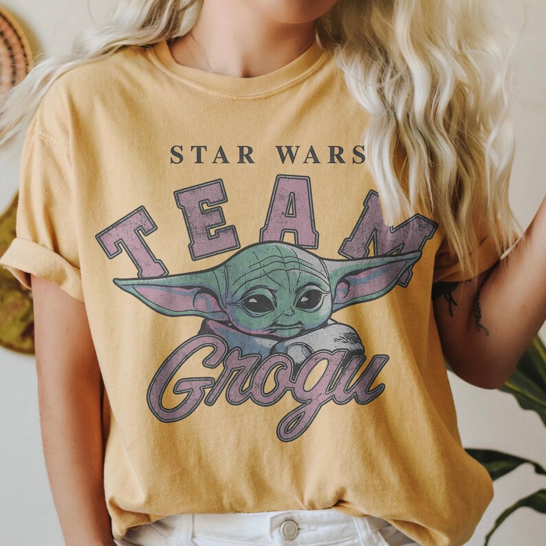 Puede incluir: Una camiseta de manga corta color amarillo mostaza con un estampado gr&aacute;fico del personaje Grogu de Star Wars. El texto STAR WARS aparece en letras negras sobre la palabra TEAM en morado y Grogu en fuente cursiva morada bajo el personaje.