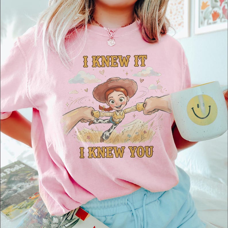 May include: A light pink t-shirt featuring a graphic of a cowgirl character with red hair and a brown hat, surrounded by clouds and flowers. The text "I KNEW IT" is printed above the character and "I KNEW YOU" is printed below in brown letters.