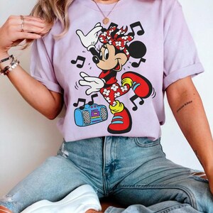 Pu&ograve; includere: Una maglietta lilla con un personaggio dei cartoni animati di Minnie Mouse che balla con note musicali. Minnie Mouse indossa un completo a pois rossi e bianchi, scarpe rosse e un fiocco abbinato. C'&egrave; anche un boombox blu nell'immagine.