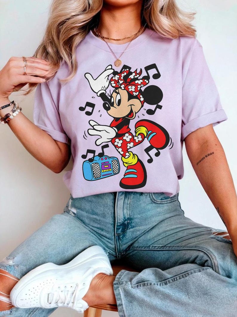 Pu&ograve; includere: Una maglietta lilla con un personaggio dei cartoni animati di Minnie Mouse che balla con note musicali. Minnie Mouse indossa un completo a pois rossi e bianchi, scarpe rosse e un fiocco abbinato. C'&egrave; anche un boombox blu nell'immagine.