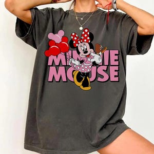 May include: Dark gray t-shirt featuring a graphic of Minnie Mouse holding balloons and a treat. Minnie Mouse is wearing a pink polka dot dress and a red bow with white polka dots. The text "MINNIE MOUSE" is in pink.