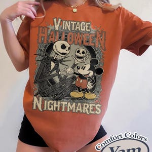May include: A burnt orange t-shirt featuring a graphic print with the text Vintage Halloween Nightmares. The design depicts a tall skeletal figure holding a skull and Mickey Mouse standing together within an ornate, dark circular frame.