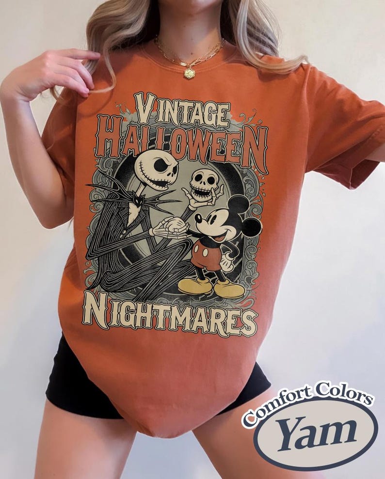 May include: A burnt orange t-shirt featuring a graphic print with the text Vintage Halloween Nightmares. The design depicts a tall skeletal figure holding a skull and Mickey Mouse standing together within an ornate, dark circular frame.