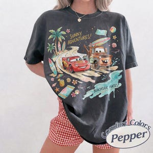 May include: A charcoal gray short-sleeve t-shirt featuring a graphic print of Lightning McQueen and Mater driving on a beach road. The text reads SUNNY ADVENTURES and BEACHSIDE MATES. The design includes palm trees, surfboards, and beach items.