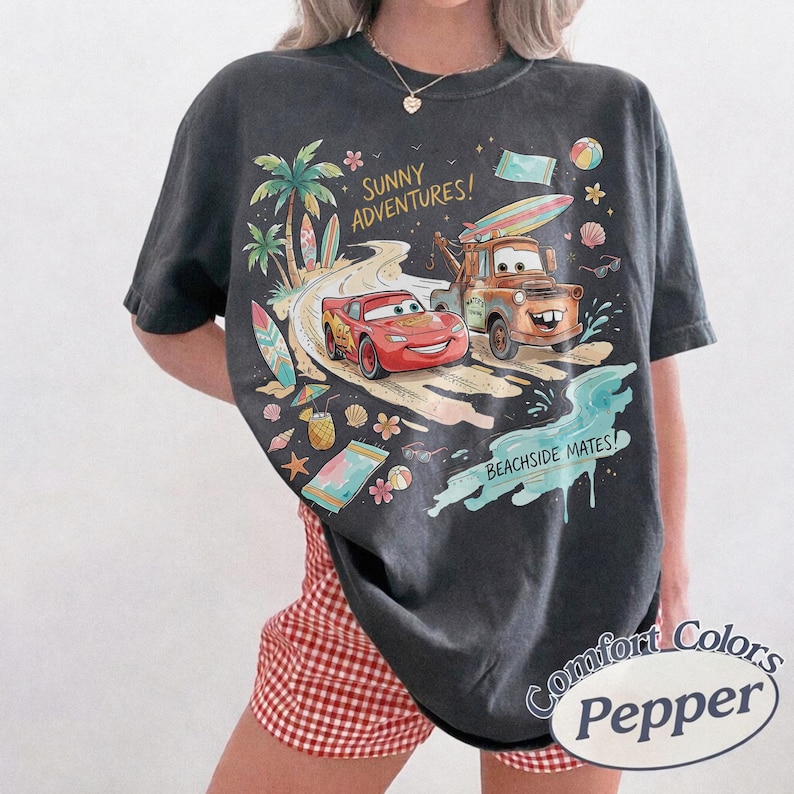 May include: A charcoal gray short-sleeve t-shirt featuring a graphic print of Lightning McQueen and Mater driving on a beach road. The text reads SUNNY ADVENTURES and BEACHSIDE MATES. The design includes palm trees, surfboards, and beach items.