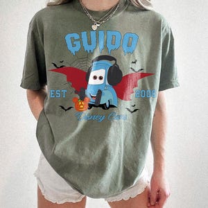 May include: An olive green t-shirt featuring a cartoon Guido from Disney Cars, with a Halloween theme. The design includes a blue Guido character with bat wings, a pumpkin, and the text "GUIDO EST 2006 Disney Cars."