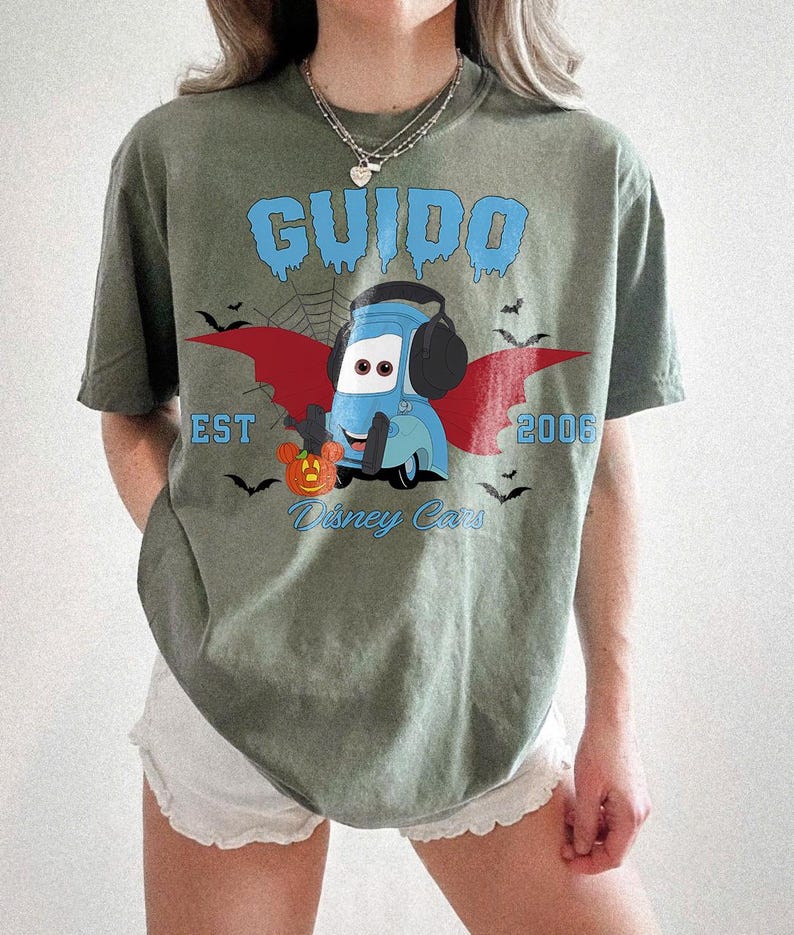 May include: An olive green t-shirt featuring a cartoon Guido from Disney Cars, with a Halloween theme. The design includes a blue Guido character with bat wings, a pumpkin, and the text "GUIDO EST 2006 Disney Cars."