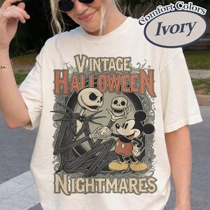 May include: An ivory t-shirt featuring a graphic print with the text Vintage Halloween Nightmares. The design depicts a tall skeletal figure in a pinstripe suit holding a skull, standing next to a cartoon mouse character in red shorts and yellow shoes.