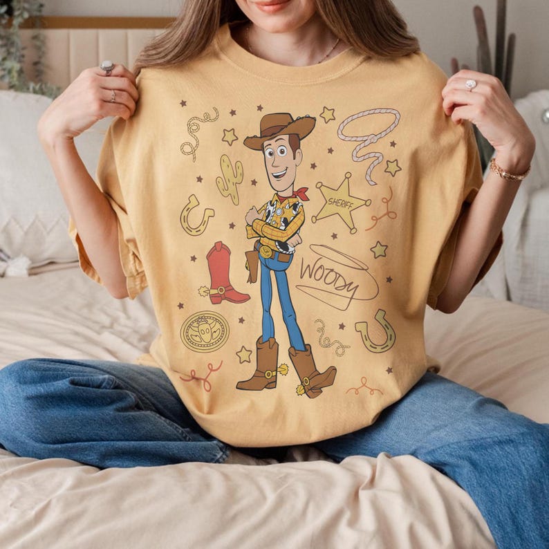 May include: Mustard yellow t-shirt featuring a cartoon illustration of Woody from Toy Story. The design includes Woody in his cowboy outfit, a sheriff's badge, a lasso, and other western-themed elements. The word "Woody" is written in a cursive font.