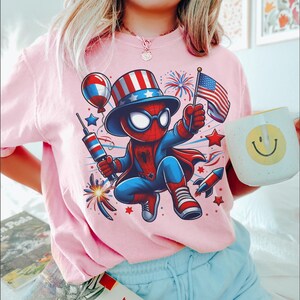 K&ouml;nnte beinhalten: Rosafarbenes T-Shirt mit einer Comicfigur von Spider-Man in einem patriotischen Outfit, die eine US-Flagge und Feuerwerk h&auml;lt. Eine wei&szlig;e Tasse mit einem gelben Smiley wird in der Hand gehalten. Das Design enth&auml;lt rote, wei&szlig;e und blaue Elemente.