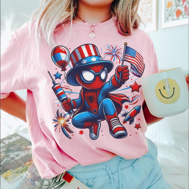 K&ouml;nnte beinhalten: Rosafarbenes T-Shirt mit einer Comicfigur von Spider-Man in einem patriotischen Outfit, die eine US-Flagge und Feuerwerk h&auml;lt. Eine wei&szlig;e Tasse mit einem gelben Smiley wird in der Hand gehalten. Das Design enth&auml;lt rote, wei&szlig;e und blaue Elemente.