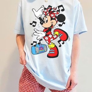 Pu&ograve; includere: T-shirt azzurra con una grafica di Minnie Mouse che balla, circondata da note musicali. Minnie indossa un completo rosso e bianco, con scarpe rosse. C'&egrave; anche un boombox blu nell'immagine. La persona indossa pantaloncini a quadri rossi e bianchi.