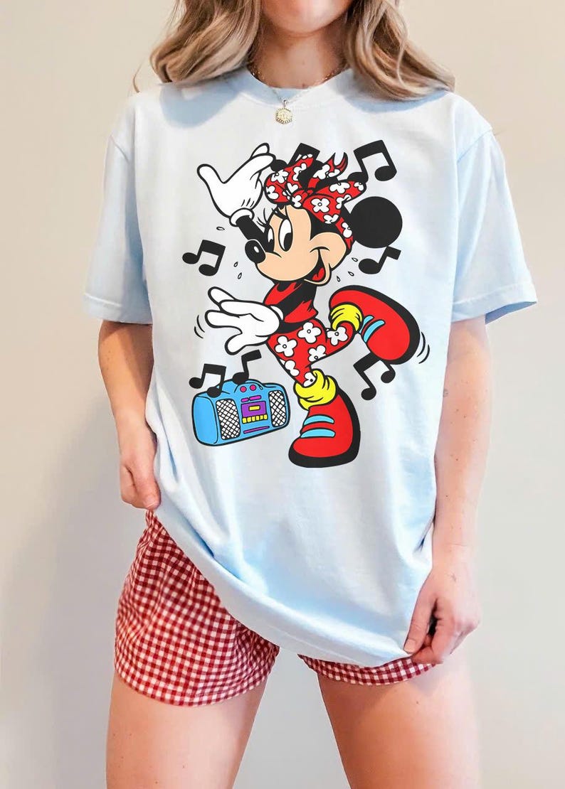 Pu&ograve; includere: T-shirt azzurra con una grafica di Minnie Mouse che balla, circondata da note musicali. Minnie indossa un completo rosso e bianco, con scarpe rosse. C'&egrave; anche un boombox blu nell'immagine. La persona indossa pantaloncini a quadri rossi e bianchi.