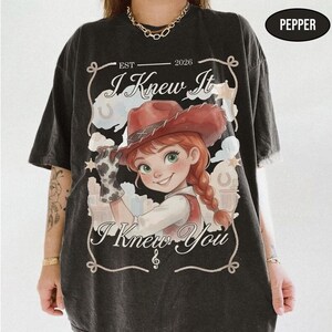 May include: A charcoal grey t-shirt featuring a graphic of a cartoon character with red hair wearing a cowgirl hat and a cow-print glove. The text reads "EST 2026", "I Knew It", and "I Knew You" within a decorative rope-style border frame.