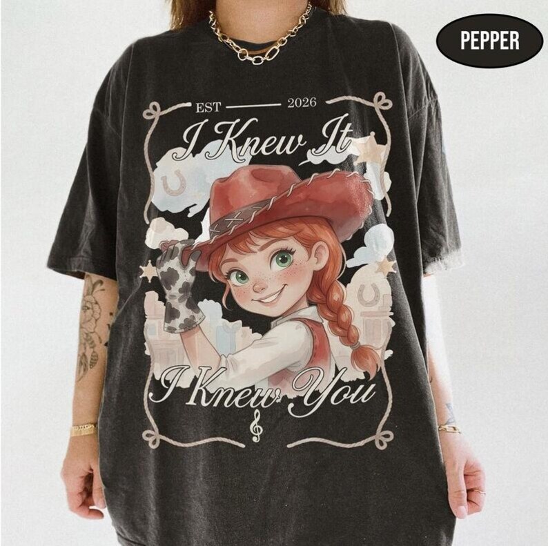 May include: A charcoal grey t-shirt featuring a graphic of a cartoon character with red hair wearing a cowgirl hat and a cow-print glove. The text reads "EST 2026", "I Knew It", and "I Knew You" within a decorative rope-style border frame.
