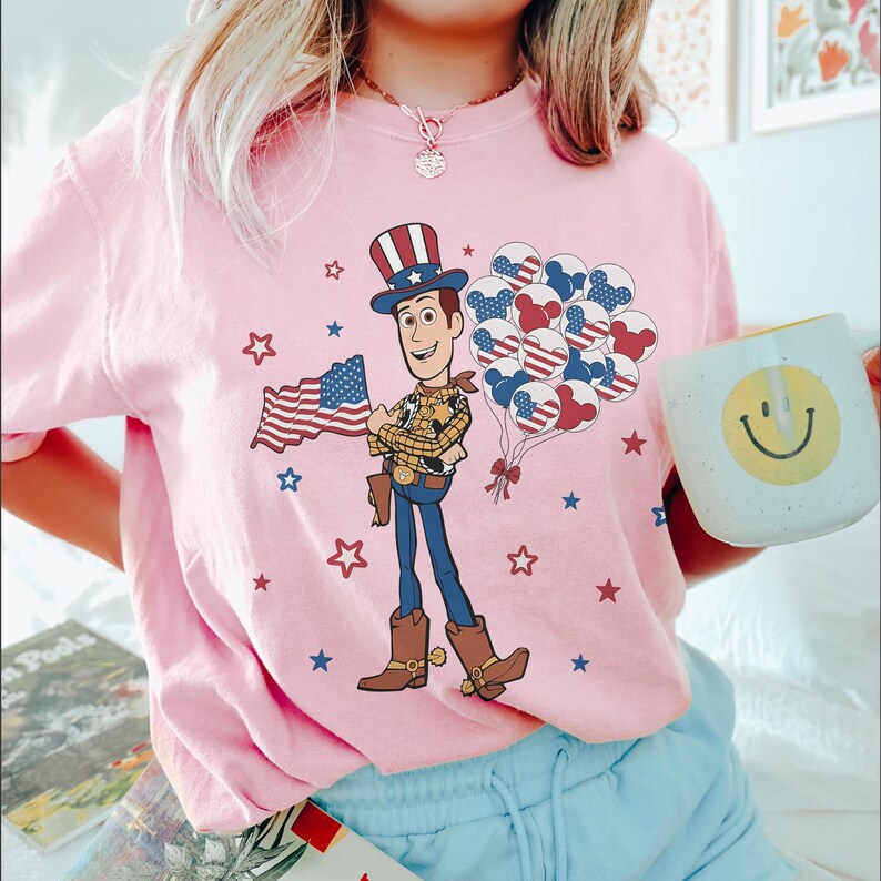 May include: Pink t-shirt with a cartoon Woody character holding an American flag and balloons. The design includes red, white, and blue stars and balloons. A white mug with a yellow smiley face is also visible.