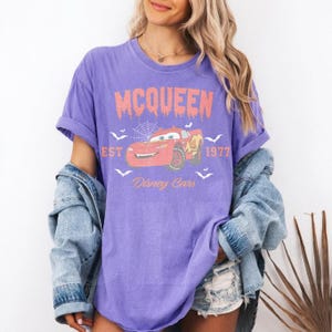 May include: A purple t-shirt featuring a graphic of Lightning McQueen from Disney Cars. The design includes the text "MCQUEEN" above the car, with "EST 1977" and "Disney Cars" below. The shirt has a vintage, distressed look.