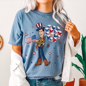 May include: A light blue t-shirt with a cartoon cowboy character holding a US flag and balloons. The balloons have a patriotic design. Red and blue stars are scattered around the graphic.