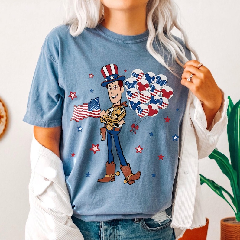 May include: A light blue t-shirt with a cartoon cowboy character holding a US flag and balloons. The balloons have a patriotic design. Red and blue stars are scattered around the graphic.