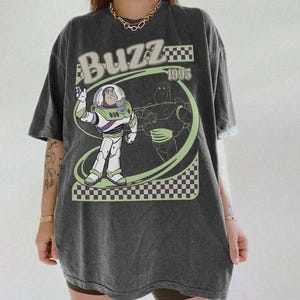 May include: Dark gray t-shirt featuring a graphic of Buzz Lightyear from Toy Story. The design includes the word "Buzz" in a light green font, the year "1995", and a checkered pattern.