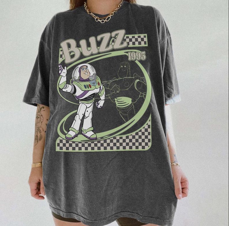 May include: Dark gray t-shirt featuring a graphic of Buzz Lightyear from Toy Story. The design includes the word "Buzz" in a light green font, the year "1995", and a checkered pattern.
