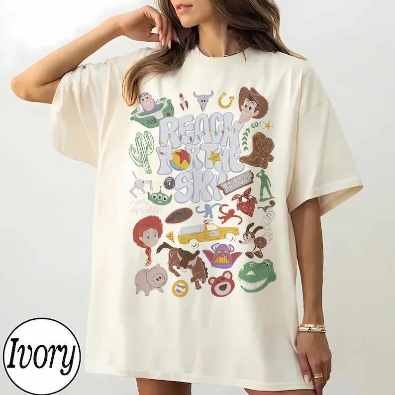 May include: An ivory t-shirt featuring a colourful graphic print with the text "REACH FOR THE SKY" surrounded by various cartoon character illustrations, including a cowboy, space ranger, green alien, pig, dinosaur, and other themed icons.